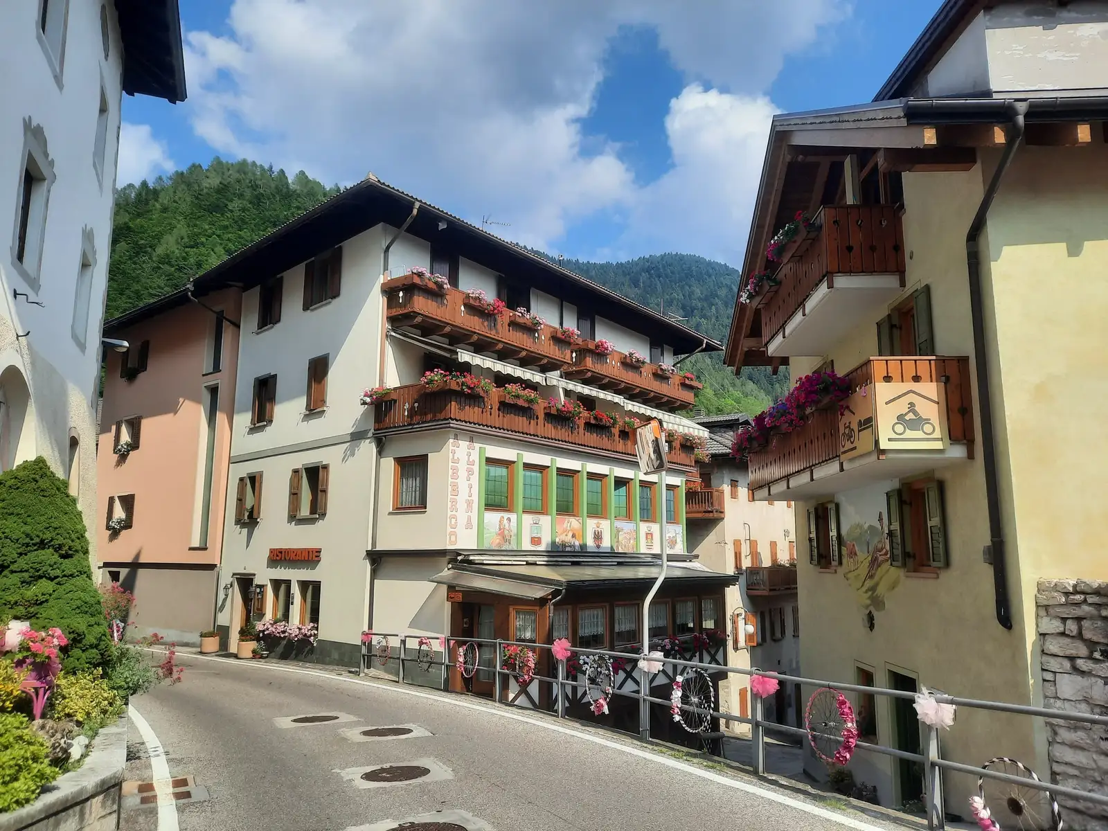 L'Albergo Alpina e la casetta di fronte lungo la via del paese, in estate, con i balconi fioriti